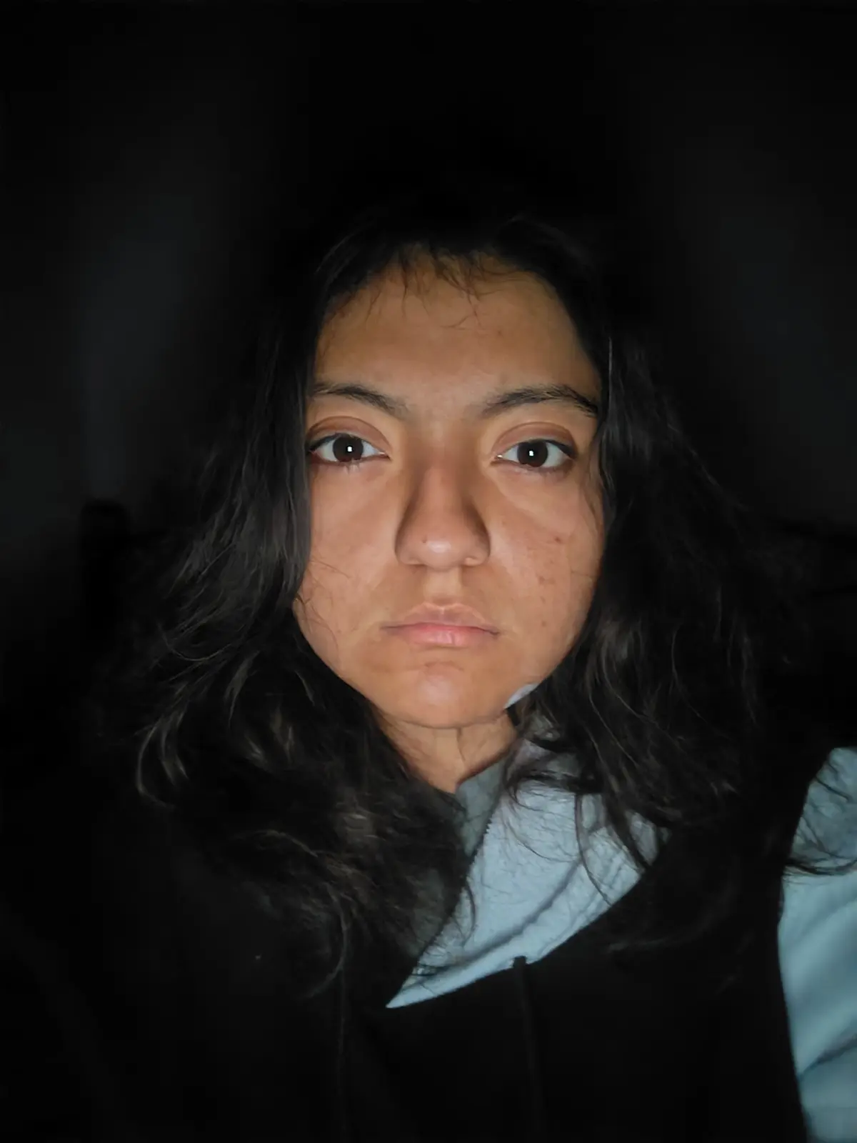 Este retrato de estudio en un plano medio corto, mirando directamente a la cámara. La paleta cromática es intencionalmente cálida y con tintes vintage: el vibrante amarillo mostaza del cuello tortuga contrasta vivamente con el verde oscuro del peto y el fondo gris azulado jaspeado, creando una separación visual que realza al sujeto. La iluminación de estudio es suave y frontal, destacando la textura del cabello y la piel, mientras que el ligero viñeteado dirige la atención ineludiblemente al centro de la composición y a esa divertida y memorable pose.