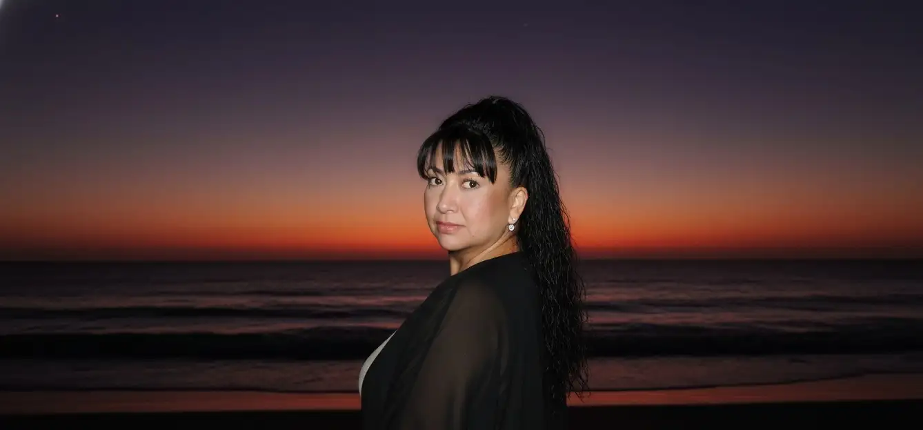 Hazme parecer que estoy en una playa oscura,

pero me sacaron una foto con flash y la cara brilla un poco. Al fondo, añade la puesta de sol, como si el sol se hubiera ocultado en un 98%, dejando un cielo rojo púrpura y casi oscuro. La imagen está tomada desde atrás y me han dicho que me gire para la foto. Llevo un traje negro playero. (Que la cara quede exactamente igual que en la foto).