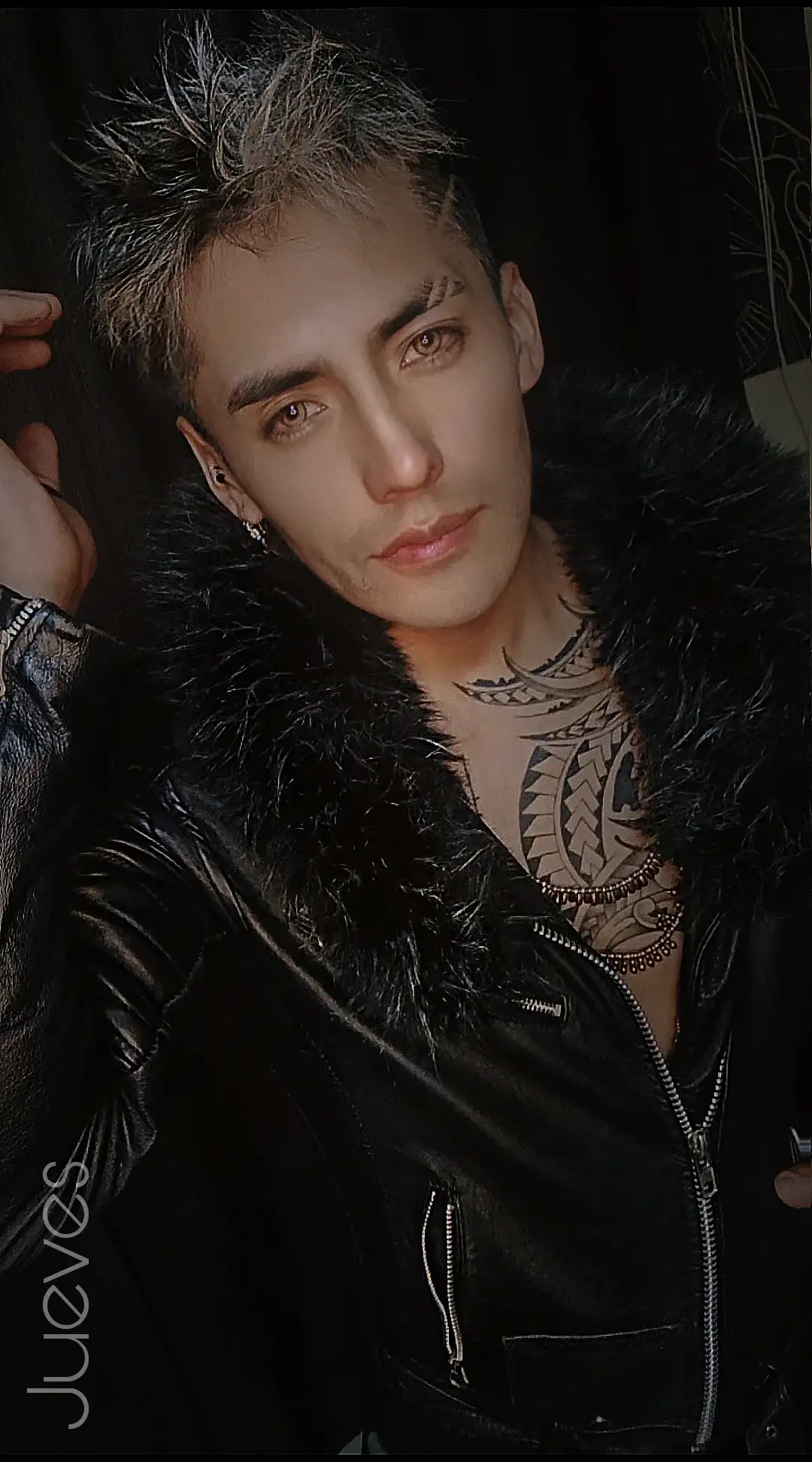 Retrato artístico y dramático de un hombre tatuado y rockero sentado en  una silla con un fondo rojo intenso iluminado con luces teatrales, viste completamente de negro con camisa blanca y con una chaqueta de cuero rojo su postura es relajada sería y poderosa cautivadora inclinado Hacia atrás con la cabeza recostada hacia atras,con los ojos cerrados transmitiendo una mezcla de intensidad vulnerable y con actitud rebelde ,el ambiente transmite sensualidad y un aire cinematográfico tomando en cuenta las características principales del rostro de la imagen adjunta