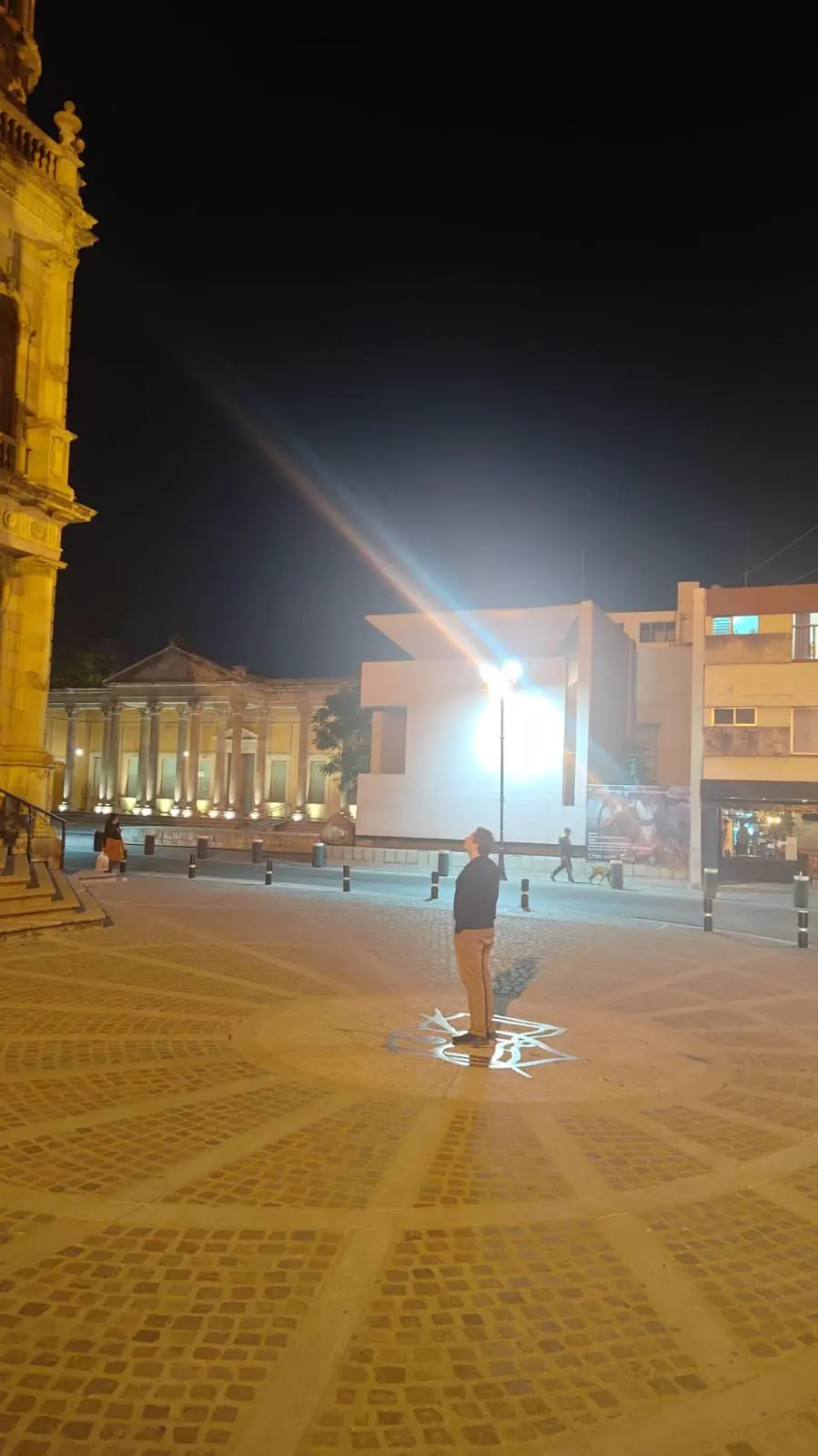Portrait-orientation, medium-long shot of Subject[Person in the Image] standing in torrential rain at night, arms flung wide with open palms, spine arched and chest lifted, head tipped back toward the downpour; low-angle 3/4 profile (~20° up). Composition: Subject centered with a slight left bias, generous headroom and surrounding negative space, diagonal rain streaks slashing across the frame as leading lines, symmetrical arm span forming strong triangular shapes, clean featureless background; crop near shins. Camera: Canon EOS 5D Mark IV (full-frame) + 50 mm, f/2.8, 1/60 s, ISO 800, white balance 3200 K; static/tripod. Lighting: single warm backlight high at 3/4 back-right creating a bright rim on jaw/arms and lighting the rain, minimal/no fill. Environment: exterior night in heavy rain with a dark background and backlit spray. Color grade: cinematic warm amber with deep blacks, moderate saturation, slightly warm temperature. Mood: cathartic, exultant, freedom. Aspect ratio 2:3. Post: S-curve contrast, clarity/micro-contrast to emphasize rain streaks, subtle vignette, mild noise reduction. Negative prompts: watermark, text artifacts, banding, oversoften skin, extra limbs, warping, blown highlights, HDR halos, plastic look.