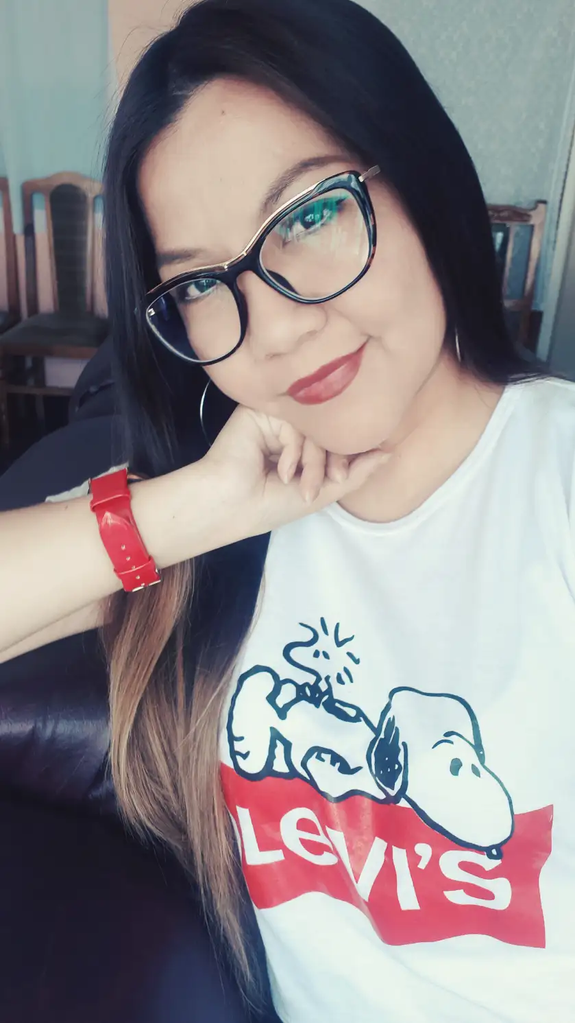 Usando mi foto, una glamurosa mujer de cabello castaño largo y liso posa con seguridad sobre un fondo de estudio neutro. Lleva un blazer negro de corte profundo sin camisa, con un estilo atrevido y elegante. Su maquillaje es impactante, con una piel impecable, delineador de ojos definido y labial rojo oscuro. Complementa su imagen con delicadas joyas de plata: un collar fino con un colgante, una pulsera y un anillo. Su expresión es feroz y segura, irradiando sofisticación y poder. Fotografía de moda profesional, alta resolución, iluminación de estudio suave, estilo editorial.