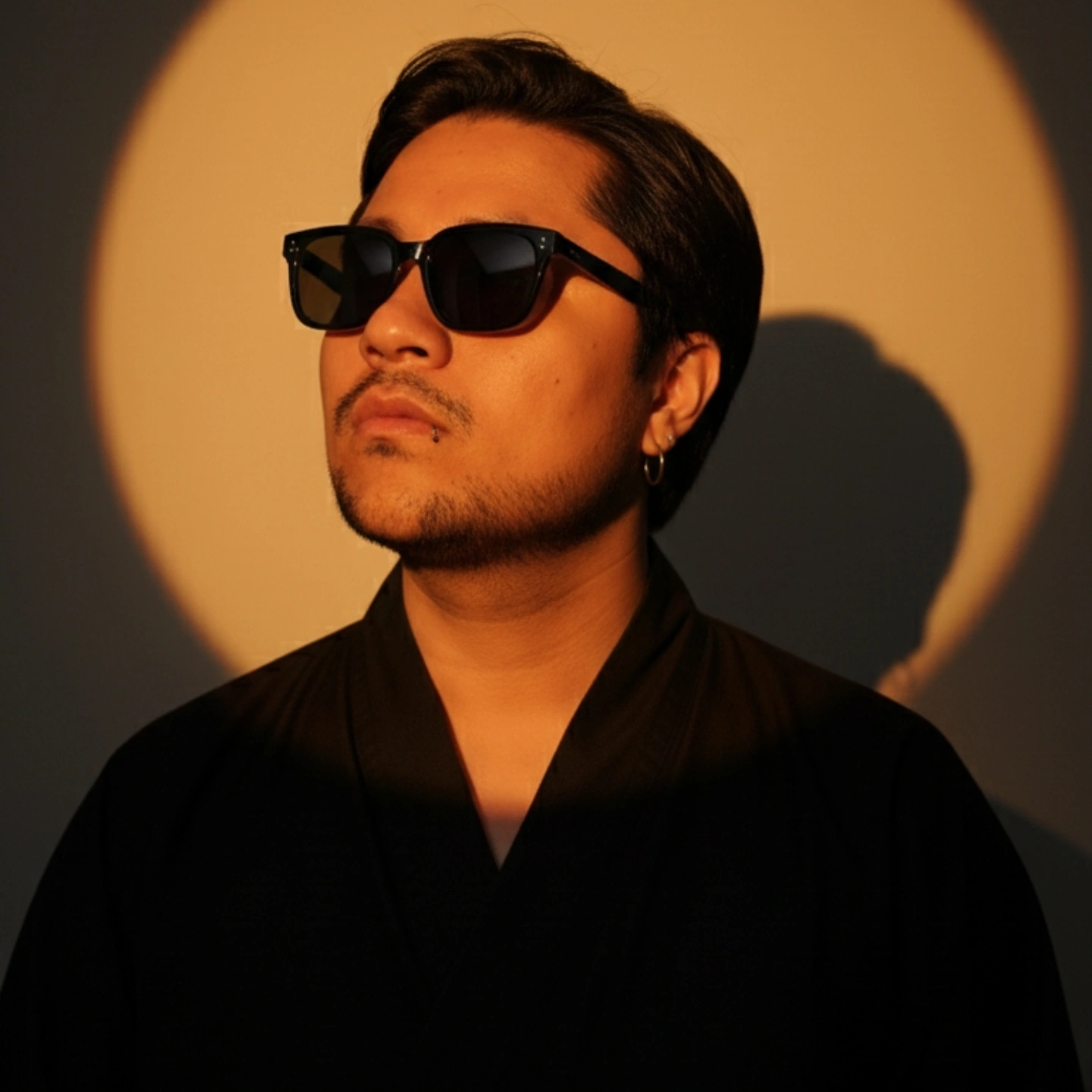 "Create a hyper-realistic professional studio portrait of the attached person, illuminated by a single golden-orange spotlight that forms a glowing circular halo behind them on the wall. Use dramatic studio lighting (Rembrandt-style, soft diffused key light with warm sunset tones) to sculpt the face and upper body, casting a strong head shadow to the right. The subject wears a black Japanese-cut shirt. Their eyes are closed, chin tilted slightly upward, expression serene. Ultra-detailed skin texture, cinematic sharpness, 85mm lens, f/1.8, full-frame DSLR quality, high dynamic range, soft background bokeh. Do not alter the facial features.