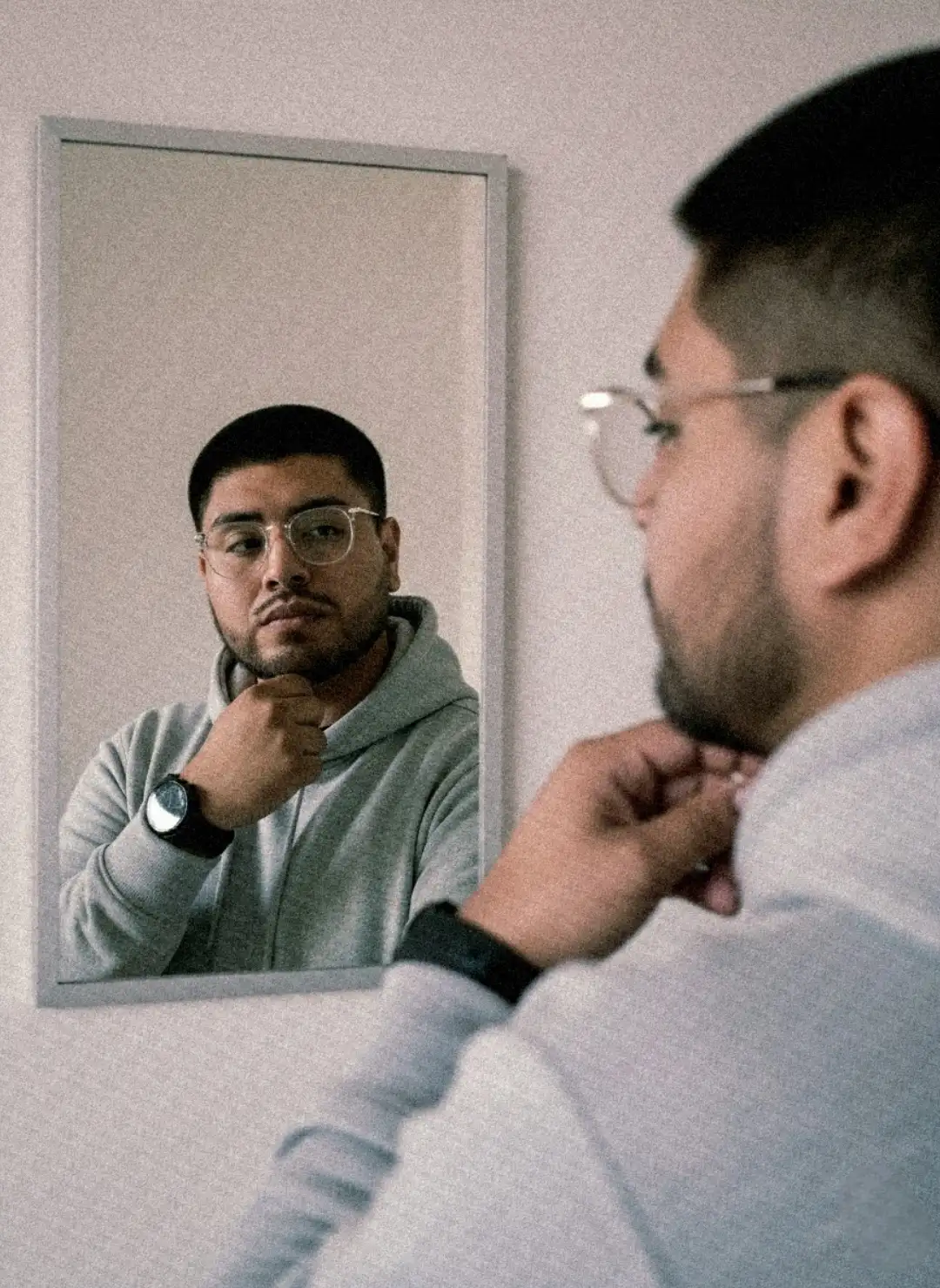 Make an image, keeping the person's facial features and face, with the following changes:
A realistic portrait photo inspired by a stylish male mirror selfie. The main focus is on the reflection in the mirror, while the real person in the foreground is softly blurred. The subject keeps his real facial and physical features, wearing glasses, a light gray hoodie (polera plomo claro) and a black wristwatch. He poses with one hand resting near his chin, gazing confidently at his reflection. Soft natural light from the side highlights the reflection’s face, creating a cinematic and modern aesthetic. The background is minimal, indoors, with neutral walls and a shallow depth of field. The overall tone is warm and slightly desaturated, shot with a portrait lens at f/2.8 using AE/AF lock, giving a professional vertical composition and ultra-realistic finish.