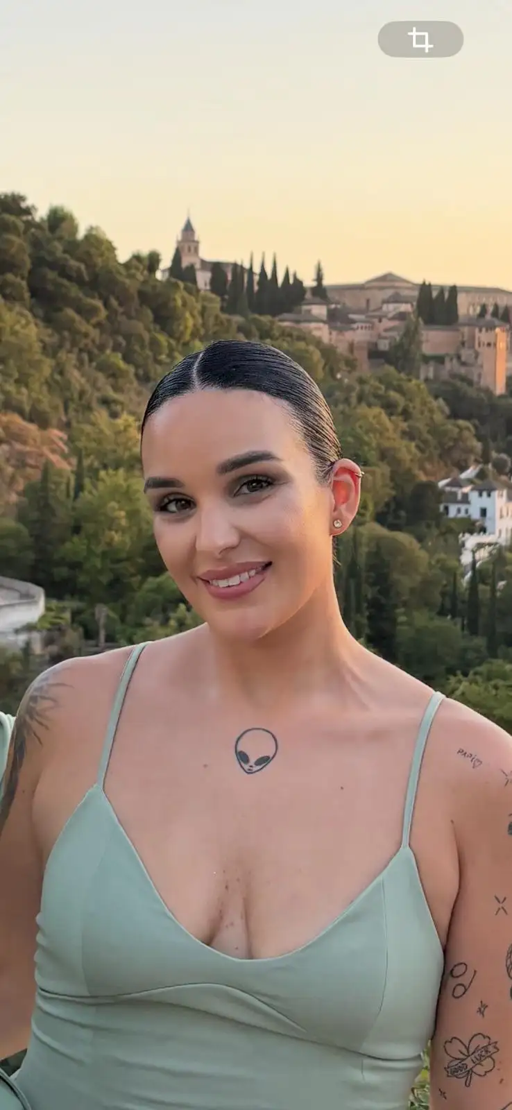 Con mi cara, crea una imagen real donde me vea más delgada, sentada en un sofá tipo taupé entero de terciopelo azul. Vestida con traje de chaqueta color crudo, la chaqueta sin abrochar, femenino y elegante y camisa de tirantes de escote recto de encaje blanca, y sandalias blancas de tacón. Manicura impecable y uña de los pies también, peinada como en la fotografía, color de pelo negro . Fondo gris con listones de madera retroiluminados y suelo de mármol gris claro